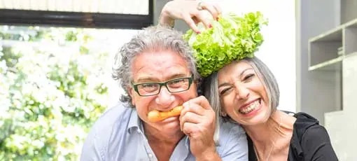 Seniorin hält sich Salatkopf auf den Kopf und Senior beisst in eine Karotte